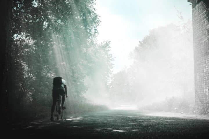 A cyclist wearing a backpack rides alone, shaded by trees.