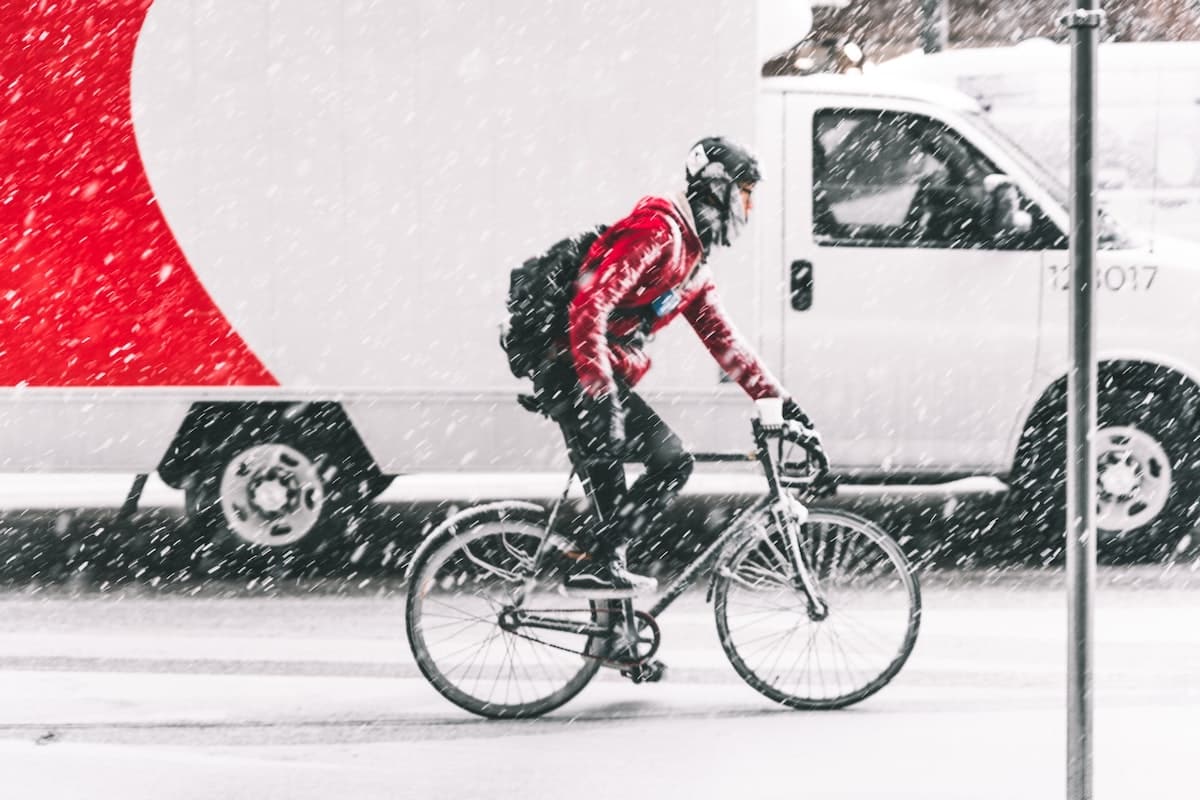 A cyclist riding in winter with snow falling on him, and a lorry driving behind him.